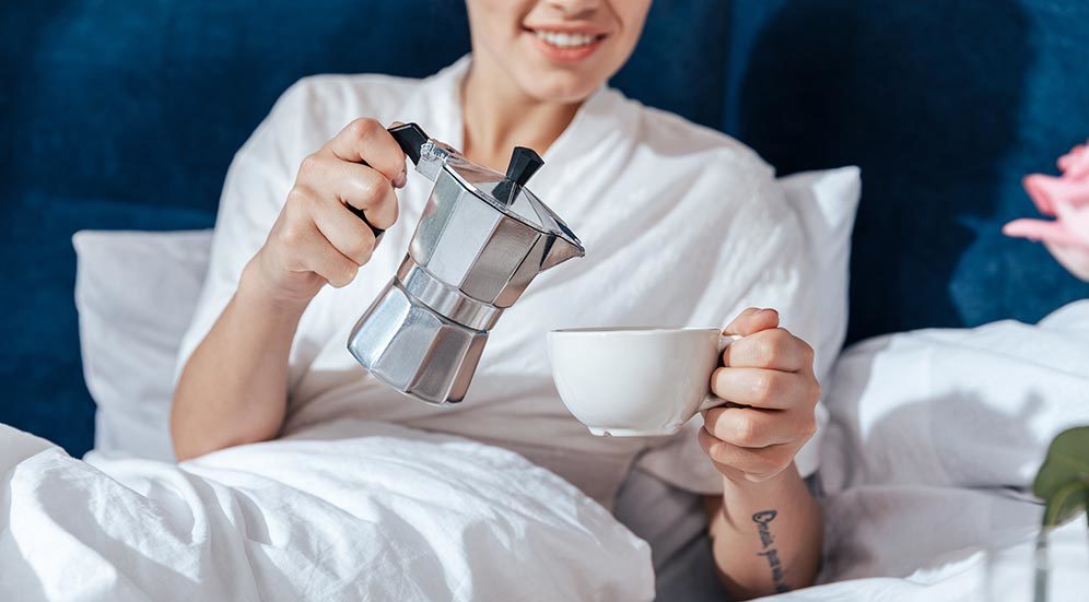 En kvinne i en seng skjenker en kopp kaffe fra en Bialetti-kanne.
