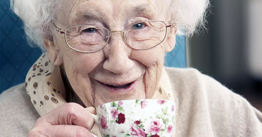 En eldre kvinne ser inn i kameraet og smiler bredt mens hun drikker kaffe