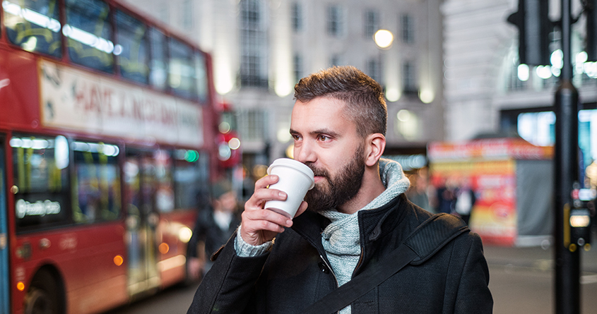 En mann drikker takeaway-kaffe i London mens en rød buss kjører i bakgrunnen.