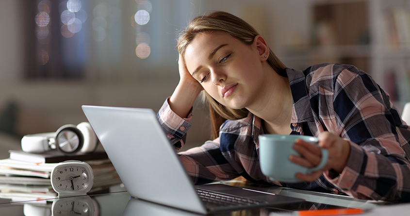 En trøtt student holder en kopp kaffe mens hun ser på dataskjermen