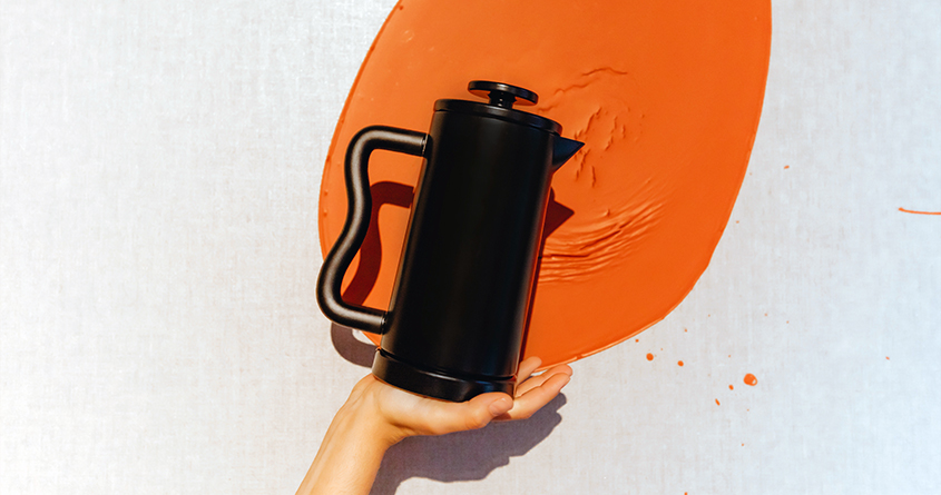En Capra Press holdes opp av en hånd foran en murvegg med en oransje malingsflekk