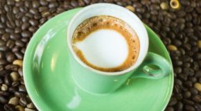 En grønn kopp oppå et lag av kaffebønner. I koppen er det en espresso med en flekk melkeskum på toppen - en espresso macchiato.