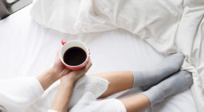 En kvinne sitter i sengen og holder en kopp med kaffe mellom hendene.