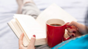 En person sitter på senga med en bok og en kopp kaffe i fanget.