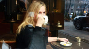 TravelQueen sitter på en kafé, hvor hun holder en kaffekopp mot munne.