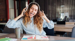 En ung kvinne sitter på kafé og smiler bredt mens hun hører på musikk og drikker kaffe