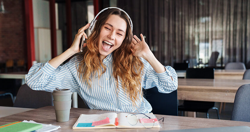 En ung kvinne sitter på kafé og smiler bredt mens hun hører på musikk og drikker kaffe