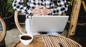 En kvinne med en laptop på fanget og en kaffe på bordet holder seg på magen sin