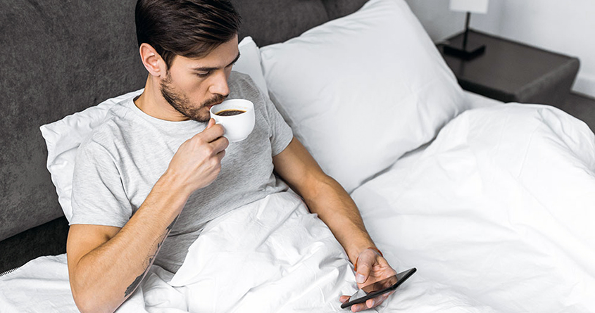 En mann sitter i senga og drikker kaffe
