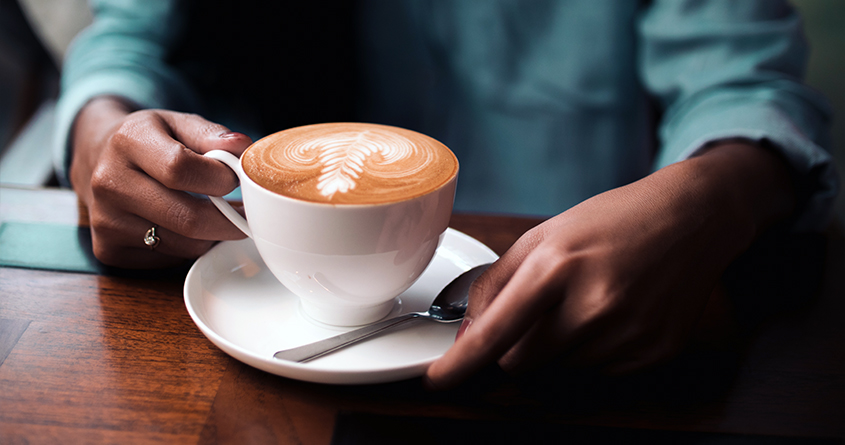 En kvinne sitter ved et bord og holder på en kopp med kaffe latte
