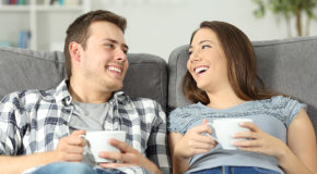 Et par sitter avslappet i en sofa og smiler mot hverandre mens de drikker kaffe.