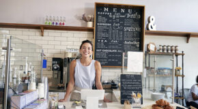 En kvinnelig barista står bak en kaffebardisk med en meny over kaffedrikker bak seg