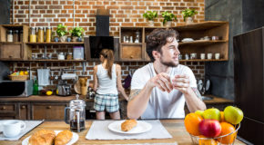 En mann sitter ved et bord og drikker kaffe mens kjæresten er i bakgrunnen.