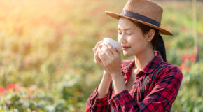 En kvinne står på en kaffefarm med lukkede øyne og nyter lukten av ferskbrygget kaffe i koppen hun holder.