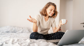En kvinne sitter i sengen og drikker kaffe mens hun videochatter.