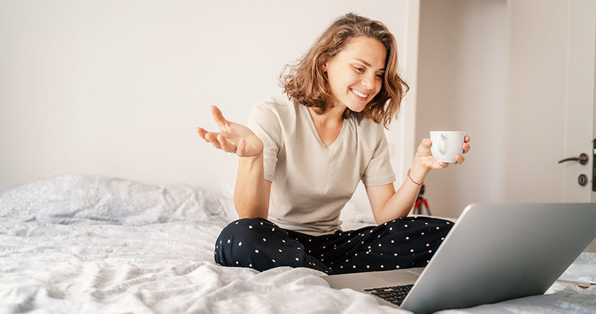 En kvinne sitter i sengen og drikker kaffe mens hun videochatter.