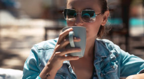 En ung kvinne med solbriller nyter en kopp med kaffe ute i solen.
