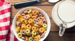 En hvit skål med fargede frokostblandingringer står på et bord, med en mugge med melk ved siden av.