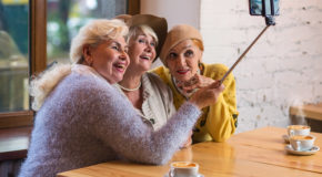 Tre eldre damer sitter på en kafé og drikker kaffe, de smiler og ler, og tar en selfie med en selfiestang.