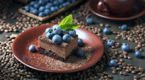 En sjokoladekake med blåbær står på et bord ved siden av en kopp med kaffe.