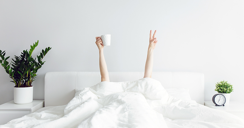 En person ligger i sengen under dynen, og strekker armene opp – én hånd viser fredstegn og den andre hånden holder en kopp med kaffe.
