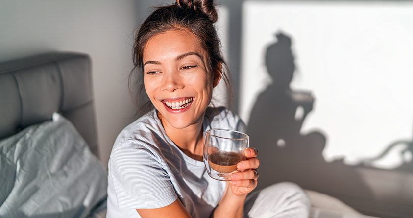 En smilende og lykkelig kvinne sitter i senga med en kopp kaffe i hånden.