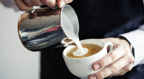 En barista lager en café con leche ved å helle melk over i en kopp med espresso.