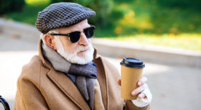 En eldre mann sitter i en rullestol ute og drikker kaffe.