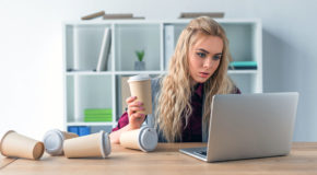 En ung kvinne sitter og arbeider for en laptop, med en kaffe i hånden og mange tomme kopper på bordet