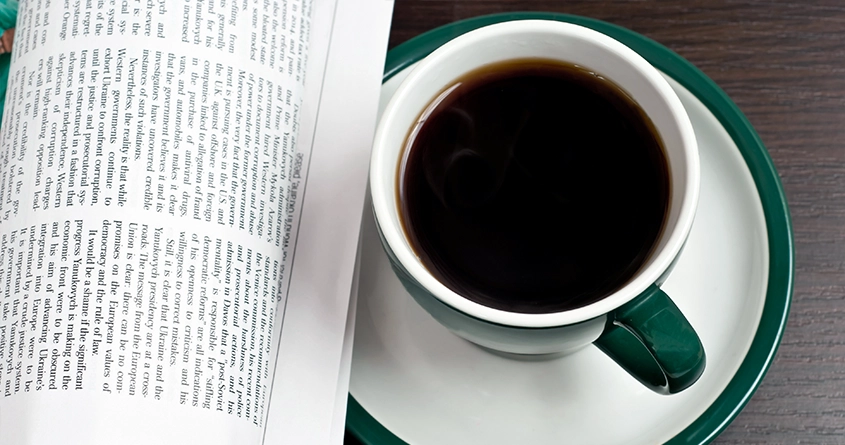 En grønn og hvit kopp med svart kaffe står på et bord ved siden av en avis