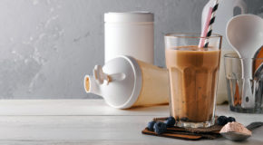 En proteinkaffe i et glass står på et bord sammen med proteinpulver og en shaker