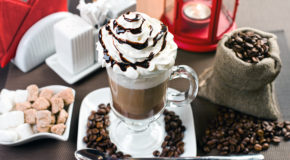 En kaffedrink med pisket krem og sjokoladesaus står på et bord sammen med kaffebønner og sukkerbiter