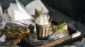 En kaffedrikk med pisket krem står på et bord ved siden av en åpen bok og noen høstblader