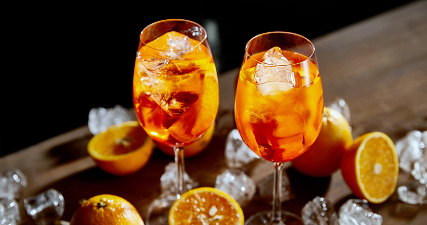 To glass med aperol spritz står på et bord, med isbiter og appelsiner liggende på bordet