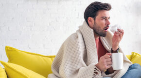 En syk mann sitter på en sofa og snyter seg med en kopp kaffe i den andre hånden