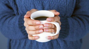 En kvinne i blå genser holder rundt en kopp kaffe med begge hender