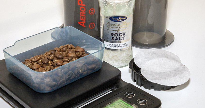Kaffebønner ligger i en skål på en vekt, ved siden av en Aeropress, to filtere og en krydderkvern