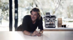 Brad Putt lener seg på en kjøkkenbenk med en kaffekopp i hånden, og med De’Longhi sin nye kaffetrakter i bakgrunnen