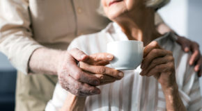En eldre kvinnen holder en kopp kaffe mens mannen hennes holder rundt henne