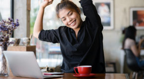 En kvinne sitter og jobber på en laptop med en kaffekopp foran seg, og smiler mens hun strekker armene over hodet
