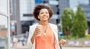 En smilende kvinne går på gaten med hodetelefoner i ørene og kaffe i hånda