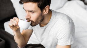 En ung mann sitter på sengekanten og drikker kaffe
