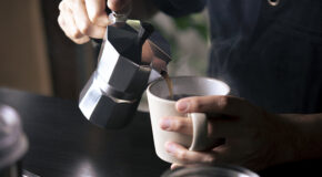 En person heller kaffe fra en perkolator oppi en hvit kopp.