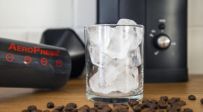 Et glass med isbiter står på en kjøkkenbenk, med en Aeropress og en kaffekvern i bakgrunnen