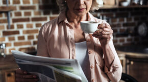 En eldre kvinne står med en kaffekopp i én hånd og leser avisen