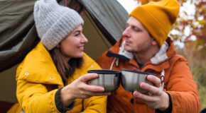 En kvinne og en mann sitter i åpningen til et telt og skåler med to kaffekopper mens de ser på hverandre