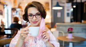 En ung kvinne sitter på kafé med en kopp kaffe i hånden og et munnbind hengende fra det ene øret.