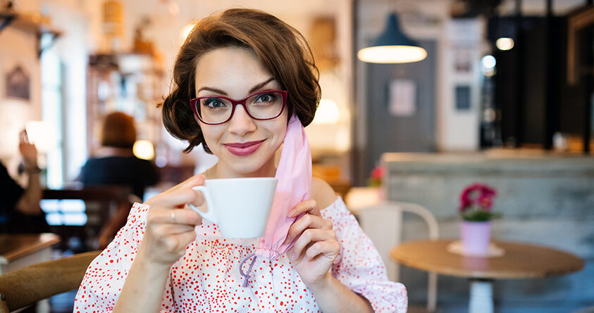 En ung kvinne sitter på kafé med en kopp kaffe i hånden og et munnbind hengende fra det ene øret.