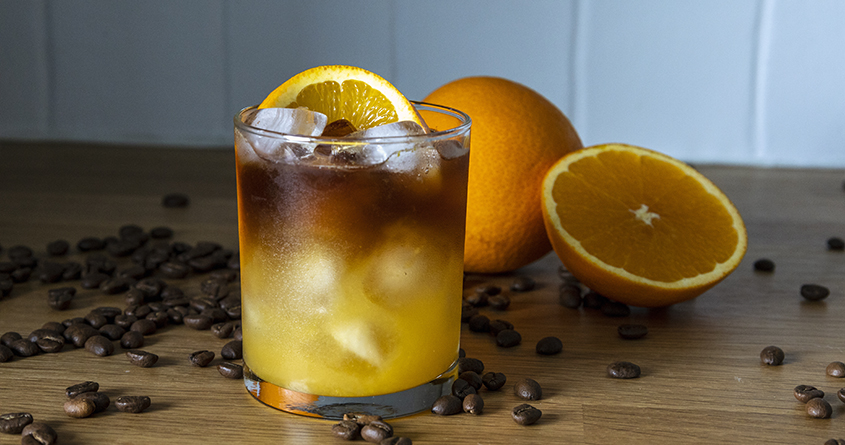 Et glass med isbiter og kaldbrygget kaffe med appelsinjuice står på en kjøkkenbenk