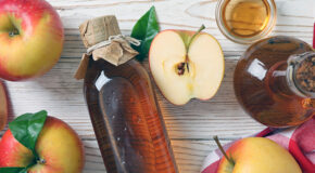 En flaske med eplecidereddik ligger på en benk ved siden av mange epler
